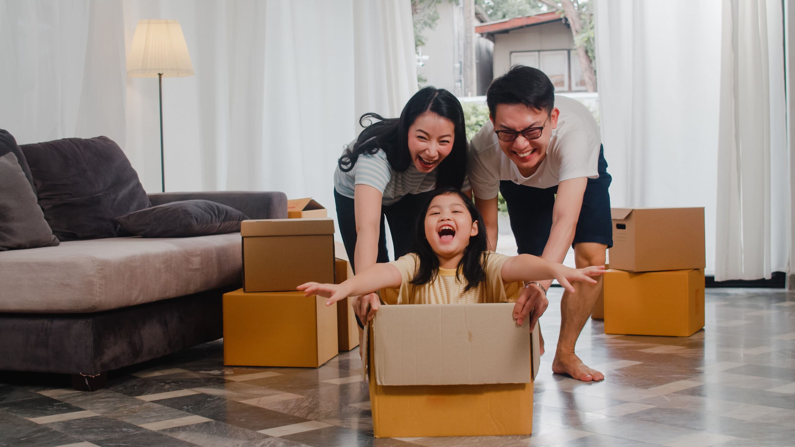 happy asian young family having fun laughing moving into new home japanese parents mother father smiling helping excited little girl riding sitting cardboard box new property relocation 1 scaled