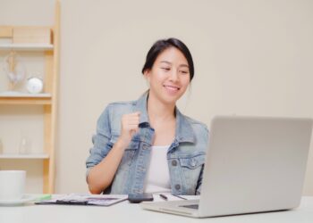 Young asian woman working using laptop on desk in living room at home. Asia business woman success celebration feeling happy dancing at home office. Enjoying time at home concept.