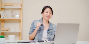 Young asian woman working using laptop on desk in living room at home. Asia business woman success celebration feeling happy dancing at home office. Enjoying time at home concept.